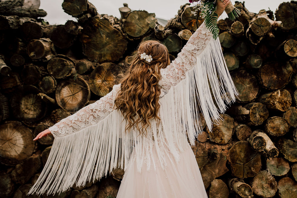 Bride facing away from the camera wearing a jacket with lots of fringing, holding up their bouquet in celebration